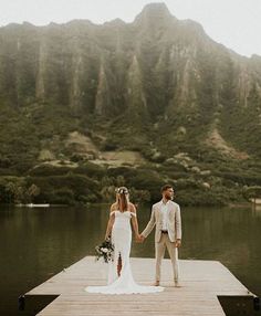 a bride and groom holding hands on a dock in front of a mountain side lake