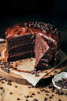 a chocolate cake with one slice cut out on a wooden cutting board next to a bowl of chocolate chips