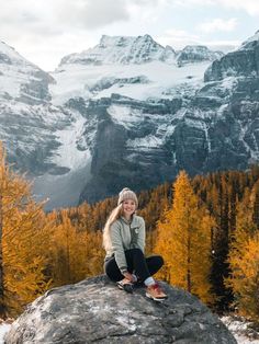 a woman sitting on top of a large rock in the middle of a forest with snow covered mountains behind her