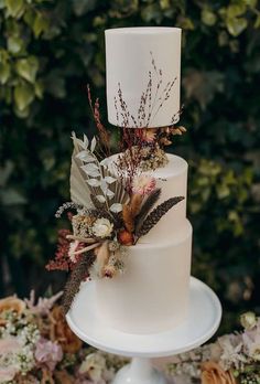 a three tiered white wedding cake with dried flowers and feathers on the top is surrounded by greenery