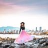 a woman in a pink dress standing on rocks near the water with a cityscape in the background