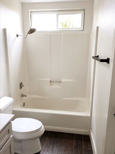 a white toilet sitting next to a bath tub in a bathroom under a window on top of a hard wood floor