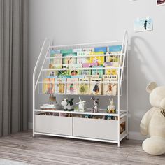 a teddy bear sitting next to a white shelf filled with books and other items on top of a hard wood floor