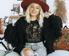 a woman sitting on a couch wearing a black shirt and fringed jacket with her hands in her hair