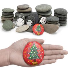 a hand is holding a rock with a christmas tree painted on it and rocks stacked in the background