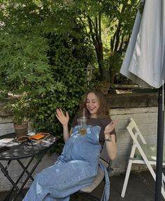 a woman in overalls sitting on a chair with her hand up to the side