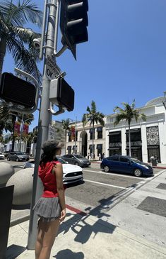 a woman standing next to a traffic light on the side of a road with palm trees and buildings in the background