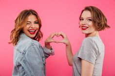 two young women making the shape of a heart with their hands against a pink background