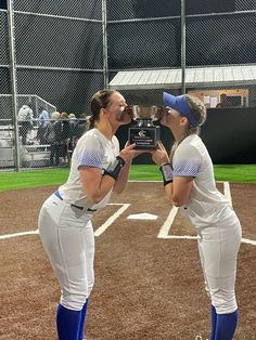 two women in white baseball uniforms standing next to each other on a baseball field holding a plaque