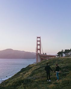 two people holding hands while walking up a hill near the water with a bridge in the background