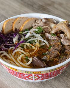 a bowl filled with meat, noodles and vegetables on top of a wooden table next to bread