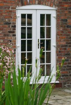 a white double door sitting next to a lush green plant in front of a brick building