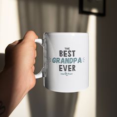 a person holding a white coffee mug with the words'best grandpa ever'on it