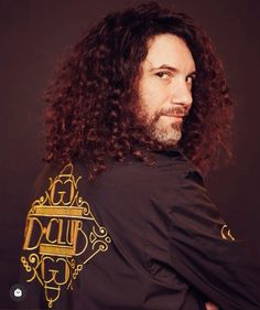 a man with long curly hair wearing a black shirt and gold lettering on his chest