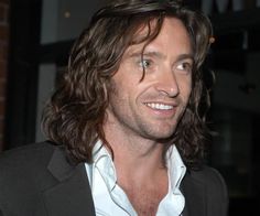 a man with long hair wearing a suit and white shirt smiles at the camera while standing in front of a brick wall