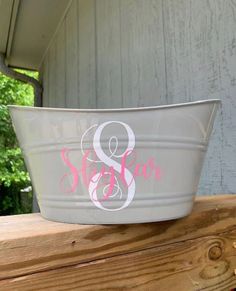 a white metal bowl with pink and gray monogrammed letters on the bottom is sitting on a wooden ledge