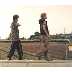 two young men walking on the side of a road next to an empty plowed field