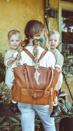 a woman carrying two small children in her back pack, both wearing braids and holding a brown purse