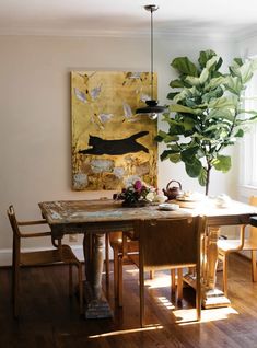 a dining room table with chairs and a large painting on the wall behind it in front of a potted plant