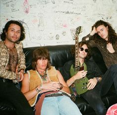 three men sitting on a couch with guitars in their hands and one man holding an electric guitar