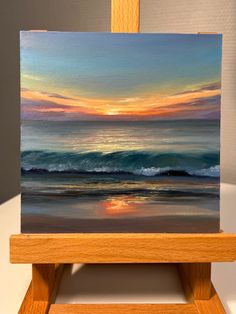 an easel with a painting on it that has a sunset in the sky and water