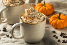 two white mugs filled with whipped cream and pumpkins on the table next to each other