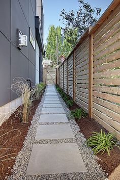 a stone path between two buildings in front of a wooden fence and planter area