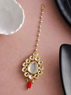 a gold necklace with white stones and red beads on a pink surface next to a flower