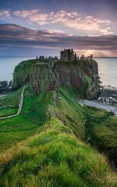 an old castle sits on the edge of a cliff overlooking the ocean at sunset with clouds in the sky