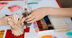 two hands holding paint sticks over a glass filled with water and other paints on a table