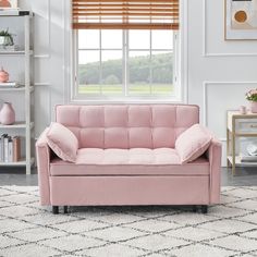 a pink couch sitting in front of a window next to a rug and bookcase