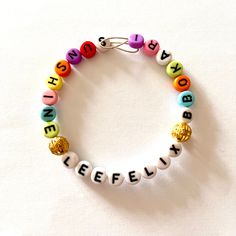 "Stray Kids Individual Member Multi-coloured Alphabet Bead Bangle - Felix. It says \"Lee Felix\" in the centre in white beads, flanked by 2 round golden filigree spacer beads and then 1 of Felix's nicknames and his SKZOO: \"Sunshine\" on the left and \"Bbokari\" on the right in multicoloured beads. The start of each flanking word is also highlighted by a white letter. *Please note that design of spacer beads will vary between bracelets, so no two bracelets will be exactly the same. The bangle it Gold Letter Beads Bracelets For Festival, Adjustable White Name Bracelet With Gold Beads, Gold Festival Bracelets With Letter Beads, Gold Bracelets With Letter Beads For Festivals, Lee Know Bracelet, Gold Beaded Bracelets With Letter Beads For Festival, White Beaded Bracelets With Letter Beads For Parties, White Beaded Bracelets With Gold Beads For Party, White Name Bracelet With Colorful Round Beads