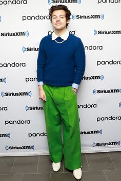 a man standing in front of a wall wearing green pants and a blue sweater with white collar