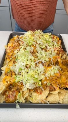 a tray full of nachos with cheese and lettuce on it next to a woman's back