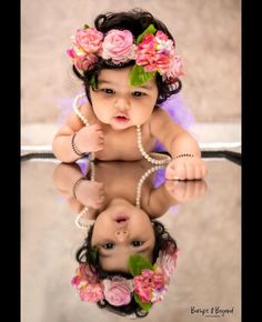 a baby girl with flowers in her hair and pearls around her neck is reflected in a mirror