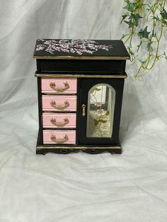 a pink and black jewelry box with flowers painted on the front, sitting next to a potted plant