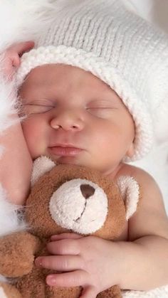 a baby sleeping next to a brown teddy bear wearing a white knitted hat and holding it's head