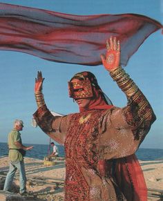 a woman dressed in red and gold on the beach with her hands raised up to catch something