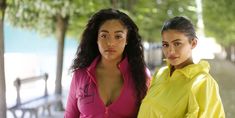 two young women standing next to each other in front of trees and water, one wearing a yellow jacket