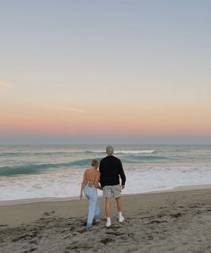 a man and woman walking on the beach at sunset with waves in the water behind them