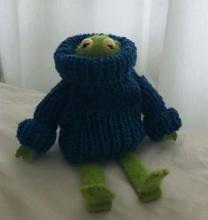 a knitted frog sitting on top of a bed next to a white curtain and window