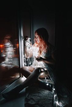 a woman sitting on a window sill looking out at the city lights in the distance