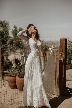 a woman standing in front of a fence wearing a white dress and holding her hands on her head