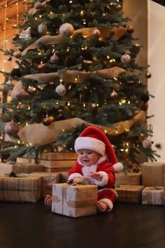 a baby sitting on the floor in front of a christmas tree with presents under it