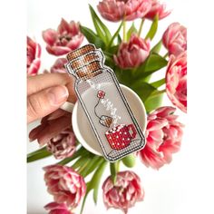 a hand holding a small bottle with some flowers in it and a cup on the side