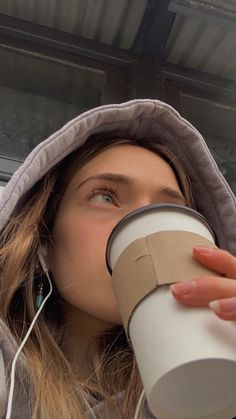 a woman drinking out of a cup while wearing earbuds and a hoodie