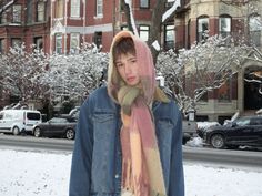 a person standing in the snow with a scarf on their head and wearing a jean jacket