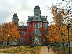 a large red building surrounded by trees with fall leaves on the ground and people walking in front