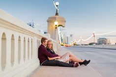 a man and woman sitting on the side of a bridge next to each other with their arms around each other