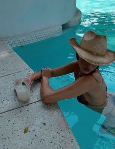 a woman in a straw hat sitting on the edge of a pool next to a drink